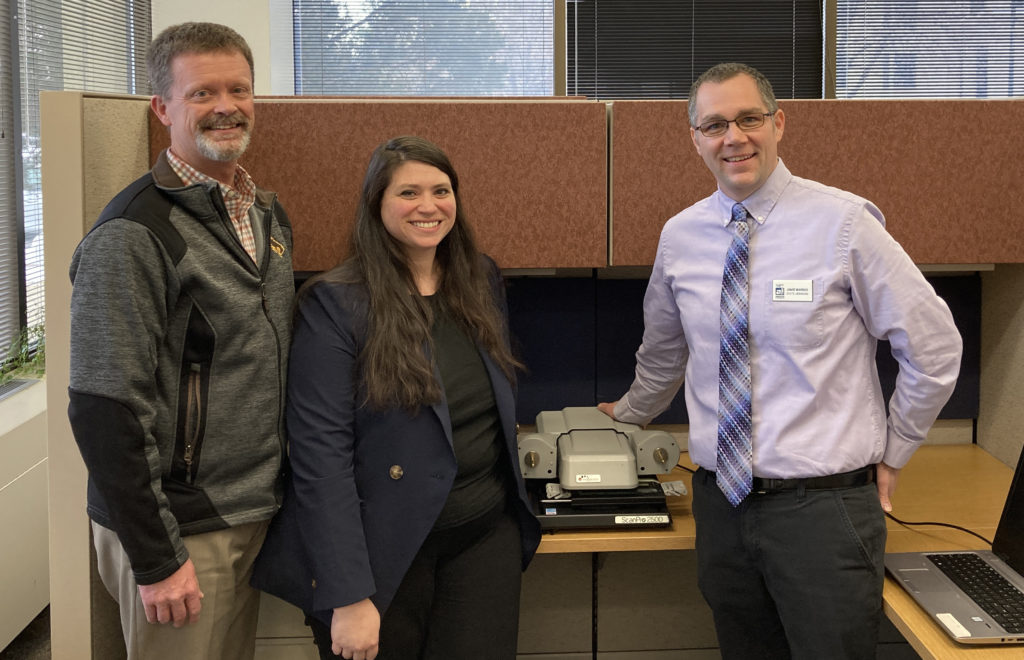 Two men and one woman standing next to microfilm scanner